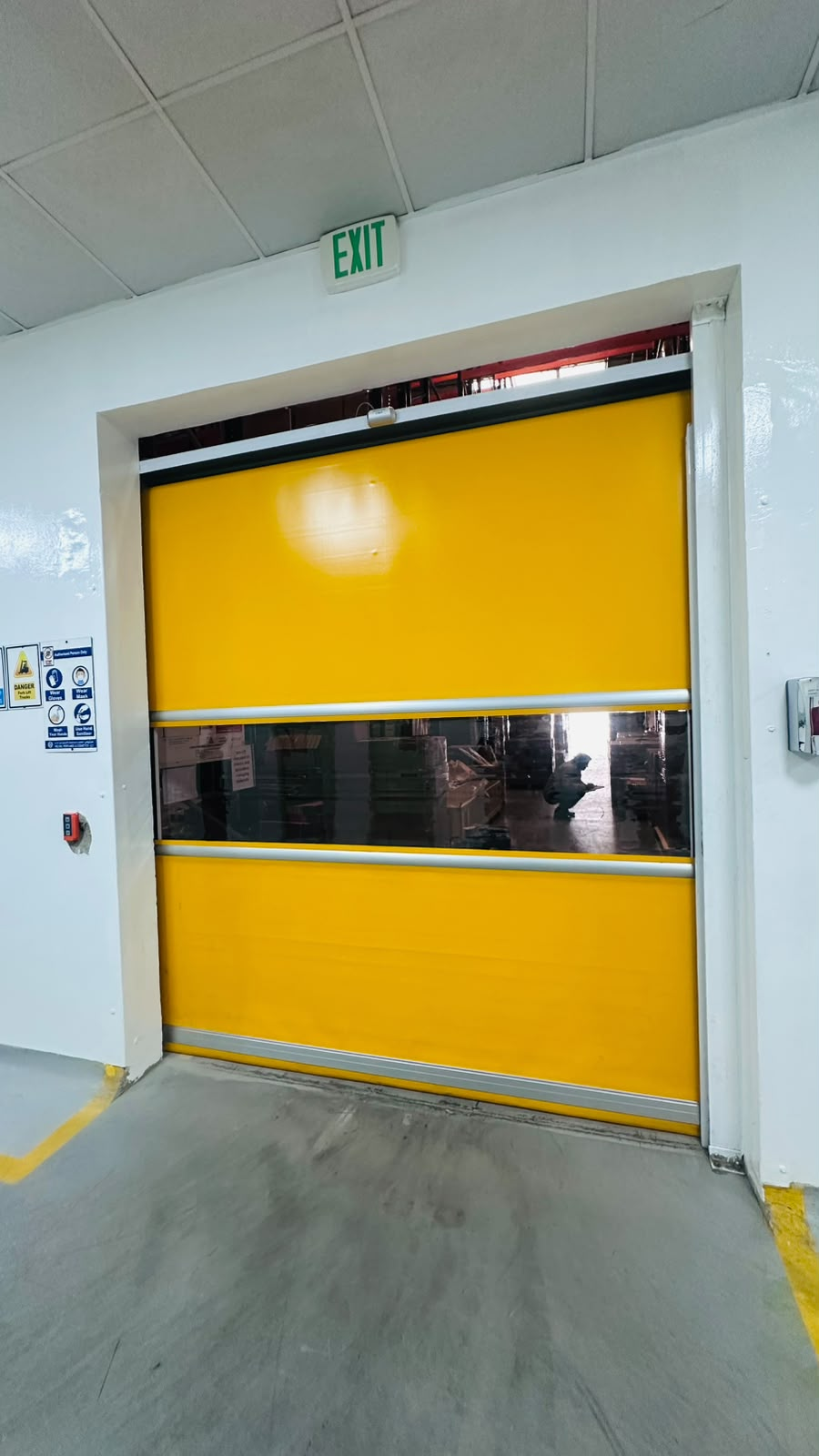 Yellow high-speed roll door with a clear vision panel fitted at an interior warehouse opening in Ras Al Khaimah, with an EXIT sign above and forklift safety signage on the wall